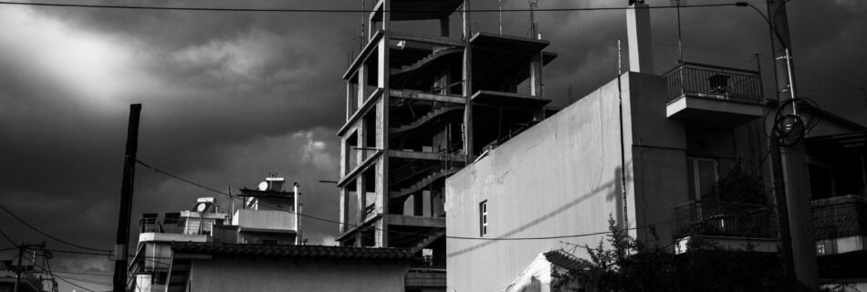 A black and white photo of an unfinished multi-story building under a dramatic, cloudy sky. In the foreground are smaller buildings, one with graffiti, and visible power lines crossing the scene.