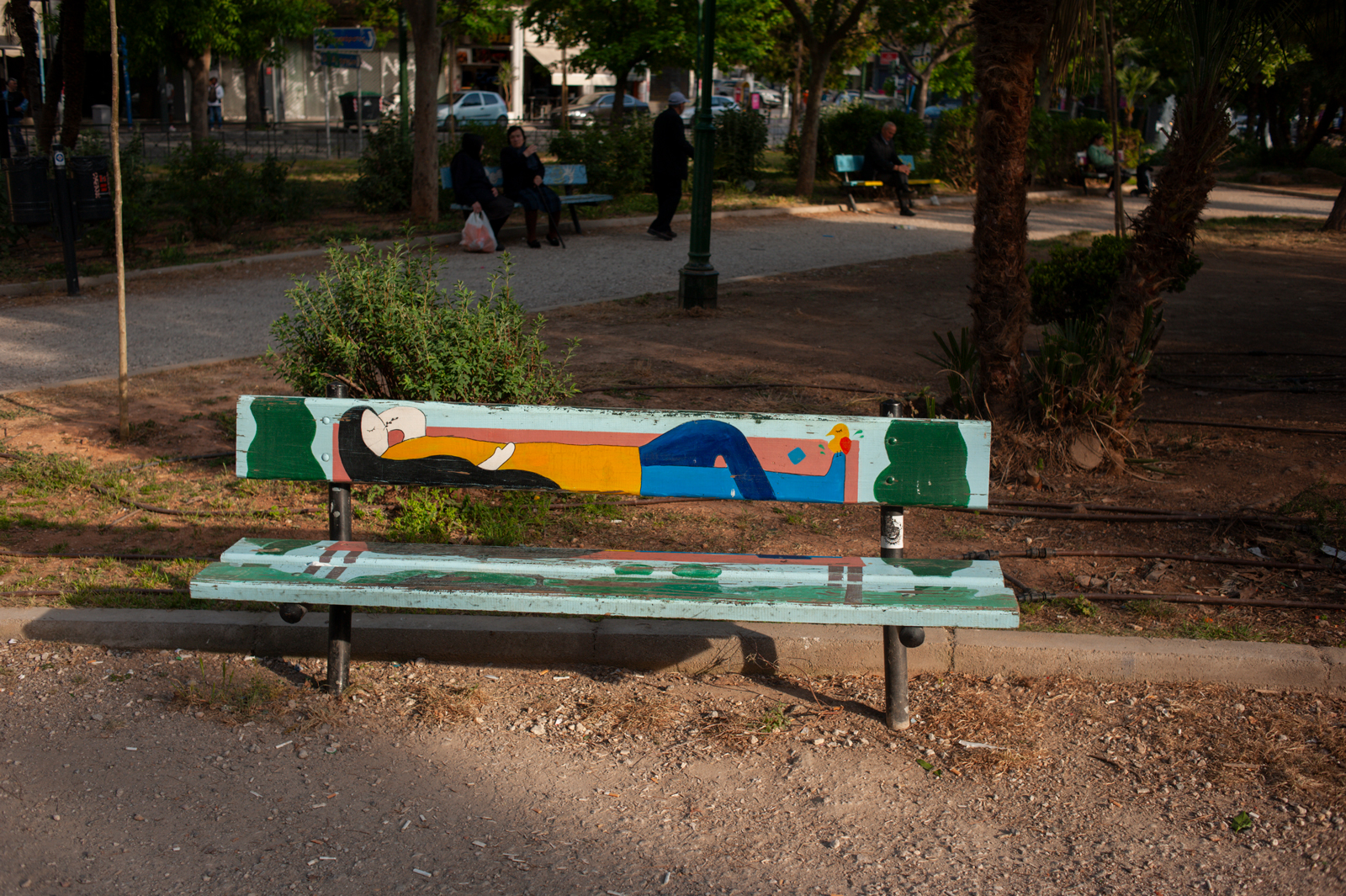 A painted park bench features a colorful, abstract image of a person lying down, set in a sunlit park with trees, paths, and people sitting on benches in the background.