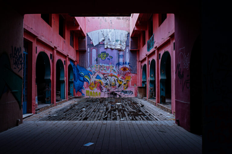 A deserted, pink-walled corridor with damaged wooden flooring, covered in colorful graffiti including animals and eyes; trash is scattered, the area appears abandoned and decayed.
