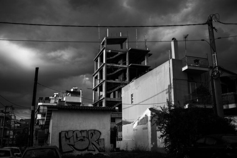 A black and white photo of an unfinished multi-story building under a dramatic, cloudy sky. In the foreground are smaller buildings, one with graffiti, and visible power lines crossing the scene.
