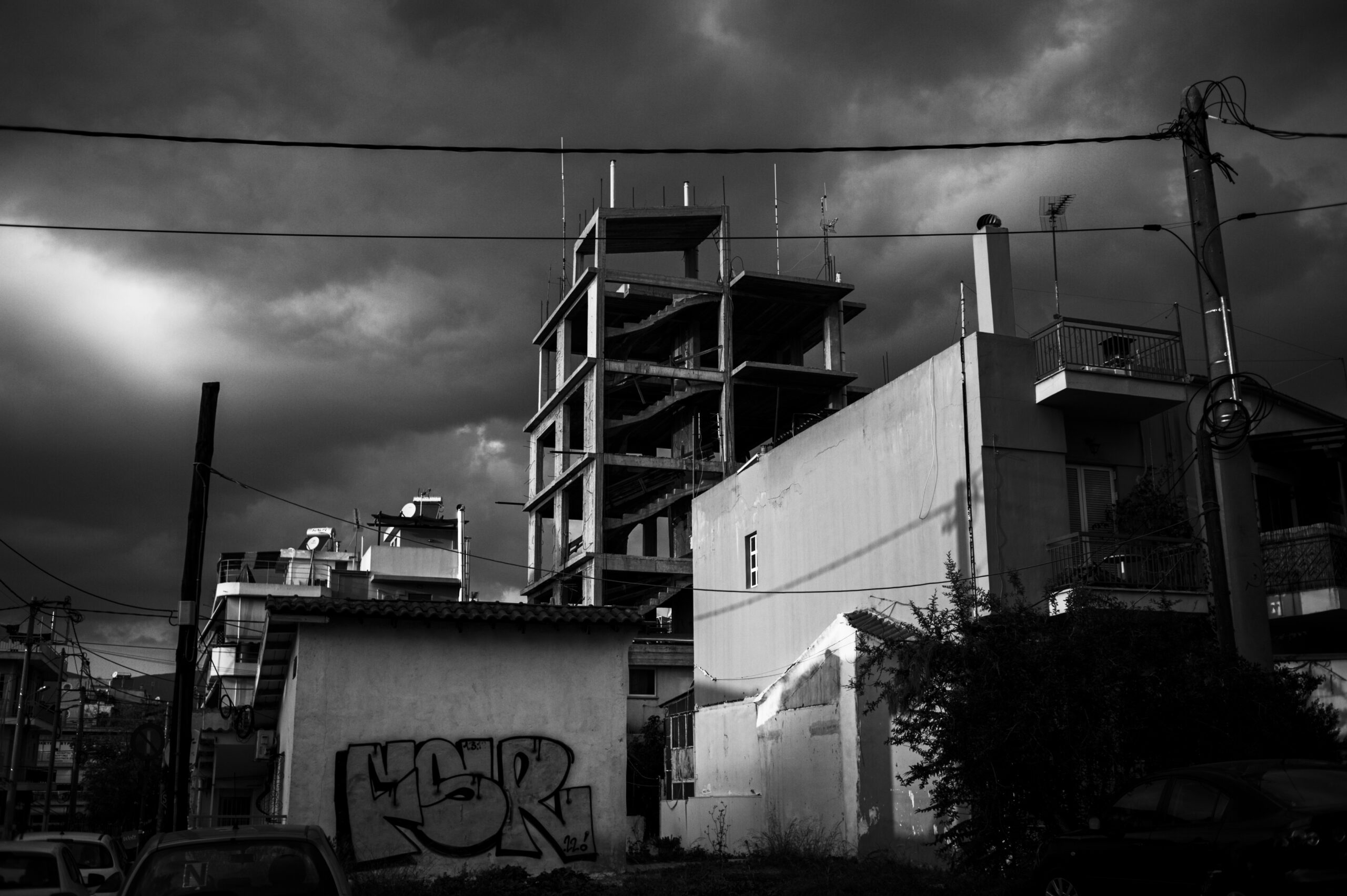 A black and white photo of an unfinished multi-story building under a dramatic, cloudy sky. In the foreground are smaller buildings, one with graffiti, and visible power lines crossing the scene.