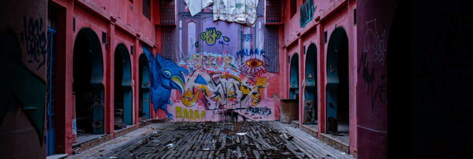 A deserted, pink-walled corridor with damaged wooden flooring, covered in colorful graffiti including animals and eyes; trash is scattered, the area appears abandoned and decayed.
