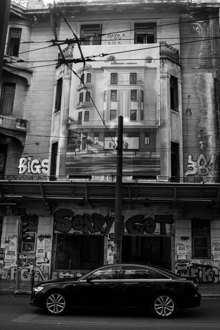 An abandoned building marked for renovation with the words "sorry I got" written on the front and a black car passing.