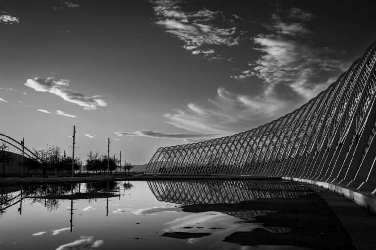 A black and white photograph of the Olympic Stadium in Athens Greece with a reflection in the water under the structure.