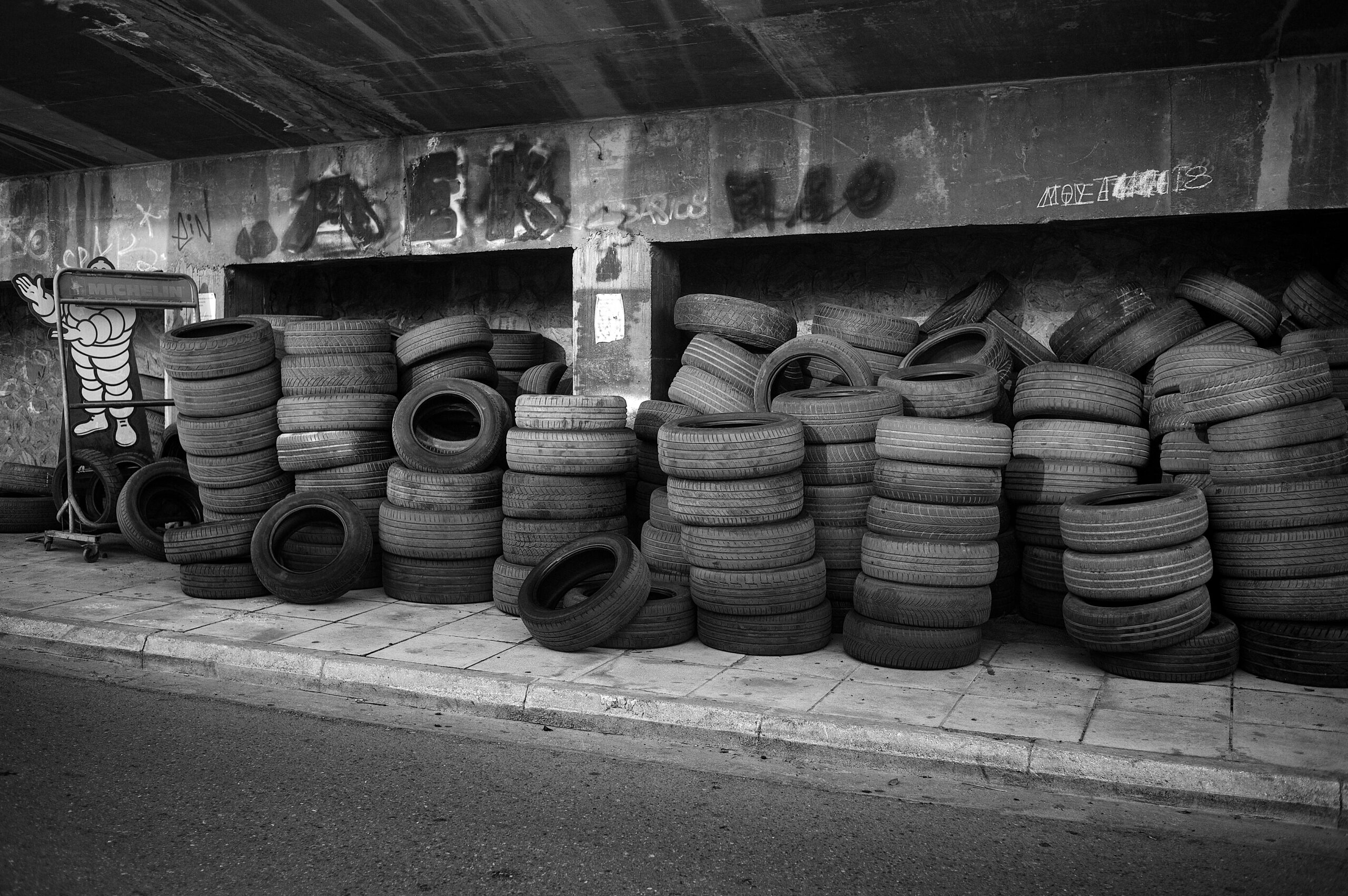 A large stack of used tires piled under a concrete overpass, with graffiti on the wall and a Michelin mascot sign to the left, captured in black and white.